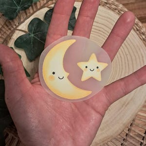Könnte beinhalten: Ein durchscheinender Aufkleber mit einem lächelnden Halbmond und einem Stern, beide mit rosigen Wangen und schwarzen Augen. Mond und Stern sind gelb mit einem hellrosa Hintergrund. Der Aufkleber wird in einer Hand gehalten.