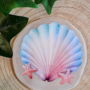 Könnte beinhalten: Dekoratives Objekt in Muschelform mit Farbverlauf in Blau, Rosa und Weiß. Zwei kleine, rosa Seesterne sind auf beiden Seiten platziert. Auf einem hellen, runden Holzsockel.