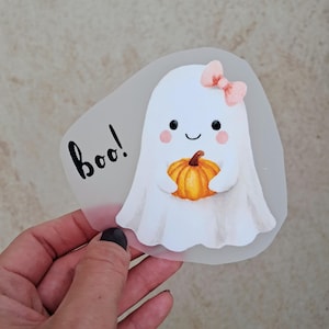 Könnte beinhalten: Illustration eines weißen Geistes mit rosa Schleife und orangefarbenem Kürbis. Das Wort "boo!" ist in schwarzer Schrift geschrieben. Das Bild hat eine durchscheinende, unregelmäßige Form, ideal für Halloween.