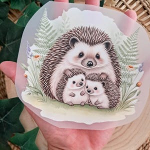 Könnte beinhalten: Ein transparenter Aufkleber mit einer Familie von Igeln. Die Illustration zeigt einen großen Igel, der zwei kleinere Igel umarmt, umgeben von Farnen und Wildblumen. Der Aufkleber hat eine weiche, pastellfarbene Farbpalette.