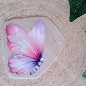 Könnte beinhalten: Illustration eines rosa und lila Schmetterlings auf einer Holzoberfläche. Der Schmetterling hat detaillierte Flügel mit Farbverläufen und zarte Fühler. Ein dekoratives Kunstwerk.