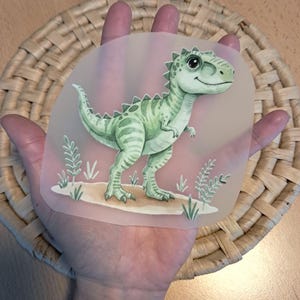 Könnte beinhalten: Illustration eines hellgrünen Dinosauriers mit freundlichem Gesichtsausdruck. Der Dinosaurier steht auf einem Sandfleck mit grünen Pflanzen. Die Illustration befindet sich auf einem klaren, abgerundeten Rechteck.