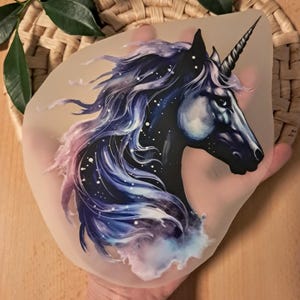 Könnte beinhalten: Detaillierte Illustration eines Einhornkopfes mit schwarzem Körper und einer fließenden Mähne in Blau-, Lila- und Rosatönen. Das Design umfasst weiße sternförmige Akzente und ein spiralförmiges Horn. Das Bild befindet sich auf einem durchscheinenden Material.