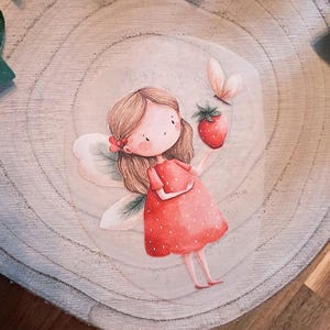 Könnte beinhalten: Eine kreisförmige Holzscheibe mit einer Illustration einer Fee mit hellbraunem Haar, die ein rotes Kleid mit weißen Punkten trägt. Die Fee hat durchscheinende Flügel, hält eine rote Erdbeere und wird von einem kleinen Schmetterling begleitet.