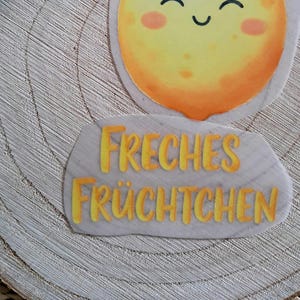 Könnte beinhalten: Eine fröhliche, cartoonartige Illustration einer lächelnden Sonne mit rosigen Wangen. Darunter steht der Text "FRECHES FRÜCHTCHEN" in Gelb auf hellgrauem, steinartigem Hintergrund. Das Bild ist vor einem Holzmaserungshintergrund platziert.