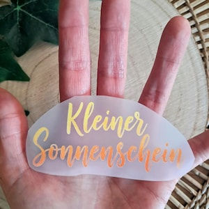 Könnte beinhalten: Ein durchscheinender, oval geformter Aufkleber mit den deutschen Worten "Kleiner Sonnenschein" in gelber und orangefarbener Schrift. Der Aufkleber wird in einer Hand gehalten, mit einem Holz- und grünen Blätterhintergrund.