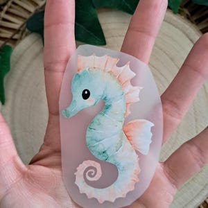 Könnte beinhalten: Ein Aufkleber mit einem Seepferdchen im Aquarellstil. Das Seepferdchen ist hellblau und grün mit pfirsichfarbenen Akzenten. Der Aufkleber hat einen transparenten Hintergrund.
