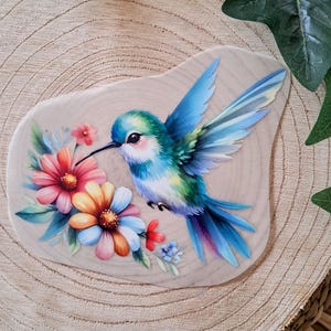 Könnte beinhalten: Eine farbenfrohe Illustration eines Kolibris mit blauen und grünen Federn, der sich in der Nähe von bunten Blumen aufhält. Das Kunstwerk befindet sich auf einem hellen Hintergrund mit Holzmaserung.