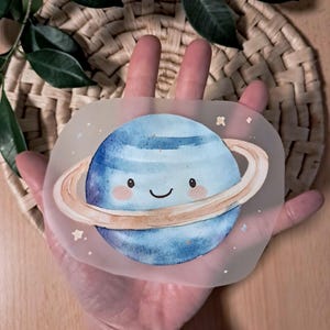 Könnte beinhalten: Ein transparenter Aufkleber mit einer Aquarellillustration eines lächelnden blauen Planeten mit Ringen. Der Planet hat ein niedliches Gesicht und ist von kleinen Sternen umgeben. Der Aufkleber wird in einer Hand gehalten.