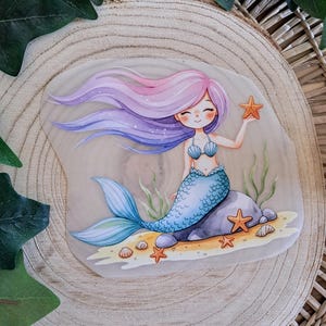 Könnte beinhalten: Eine skurrile Illustration einer Meerjungfrau mit fließendem lila und rosa Haar, einem blauen Muscheloberteil und einem blauen Schwanz. Sie hält einen orangefarbenen Seestern, mit anderen Seesternen und Muscheln auf dem sandigen Meeresboden. Das Kunstwerk ist vor einem hellen Hintergrund.