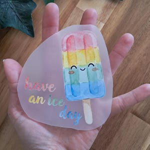 Könnte beinhalten: Ein transparenter Aufkleber mit einem lächelnden, regenbogenfarbenen Eis am Stiel-Design. Der Text "have an ice day" ist in einer verspielten Schriftart geschrieben. Der Aufkleber wird in einer Hand gehalten. Das Eis hat einen Holzstiel.