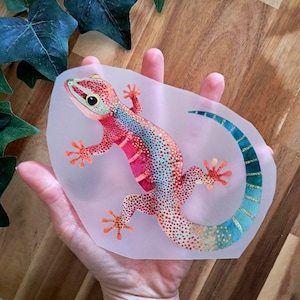 Könnte beinhalten: Bunte Gecko-Illustration in Rot-, Rosa-, Blau- und Orangetönen. Der Gecko hat ein detailliertes Muster und ist vor einem durchscheinenden Hintergrund platziert. Das Bild wird in einer Hand gehalten, mit grünen Efeublättern im Hintergrund.
