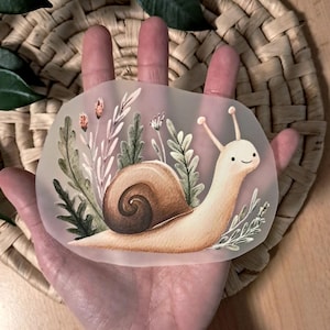 Könnte beinhalten: Ein durchscheinender Aufkleber mit einer Illustration einer Schnecke. Die Schnecke hat ein braunes Gehäuse und ein lächelndes Gesicht, umgeben von grünen und rosa floralen Elementen. Der Aufkleber wird in einer Hand gehalten.