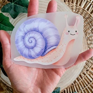 Könnte beinhalten: Ein skurriler Aufkleber mit einer Cartoon-Schnecke mit einem lila-blauen Gehäuse. Die Schnecke hat einen rosa Körper mit kleinen Punkten und einem lächelnden Gesicht. Der Aufkleber wird in einer Hand gehalten.