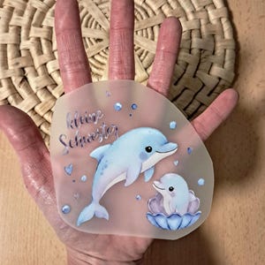Könnte beinhalten: Ein transparenter Aufkleber mit einem hellblauen Delphin und einem Baby-Delphin in einer Muschel. Der Aufkleber trägt den deutschen Text "kleine Schwester" und ist mit blauen Blasen und Herzen verziert.