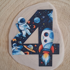 Könnte beinhalten: Eine blaue und schwarze Zahl vier mit Weltraumthema. Die Zahl ist mit einem Astronauten, einer Rakete, Planeten und Sternen verziert.