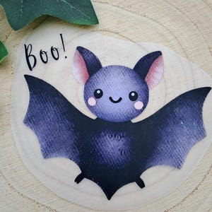 Könnte beinhalten: Ein niedlicher Cartoon-Fledermaus mit einem lächelnden Gesicht und ausgebreiteten Flügeln. Die Fledermaus ist hauptsächlich dunkelviolett mit helleren violetten Ohren und rosa Wangen. Das Wort "Boo!" steht in schwarzer Schrift über der Fledermaus. Auf einer hellen Holzoberfläche.