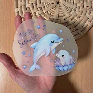 Könnte beinhalten: Ein durchscheinender Aufkleber mit zwei Cartoon-Delfinen in Blautönen. Ein Delfin schwimmt, der andere ruht in einer blauen Muschel. Der geschwungene Text lautet "große Schwester".