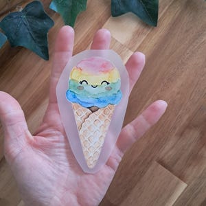 Könnte beinhalten: Ein Aufkleber in Form einer lächelnden Eistüte mit einer bunten Eiskugel. Die Eiskugel hat Regenbogenfarben. Der Waffelkegel ist hellbraun. Der Aufkleber wird in einer Hand gehalten.