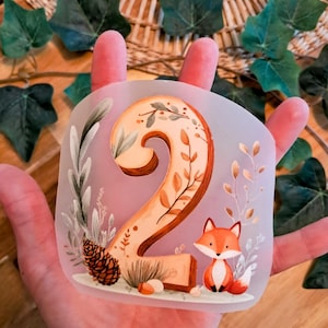 Könnte beinhalten: Eine durchscheinende, halbflexible Silikonform mit einer erhabenen Nummer zwei. Die Zahl ist mit botanischen Illustrationen von Blättern und Zweigen verziert, begleitet von einem Tannenzapfen und einer kleinen Fuchsgrafik an der Basis.