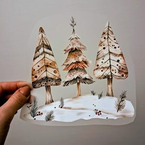 Könnte beinhalten: Aquarellillustration von drei stilisierten Weihnachtsbäumen in Braun-, Beige- und Weißtönen. Die Bäume stehen vor einer schneebedeckten Landschaft mit roten Beeren und Tannenzweigen. Das Bild wird von einer Hand gehalten.
