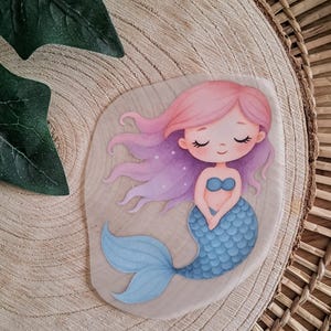 Könnte beinhalten: Eine skurrile Illustration einer Meerjungfrau mit rosa Haaren und blauem Schwanz. Die Meerjungfrau hat geschlossene Augen und ein sanftes Lächeln und trägt ein blaues Muscheloberteil. Das Kunstwerk ist vor einem hellen Holzmaserungshintergrund platziert.