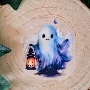 Könnte beinhalten: Ein Aufkleber mit einem freundlichen Geist in Blau- und Lilatönen, der eine Laterne mit einem geschnitzten Kürbisgesicht hält. Im Hintergrund ist ein Halbmond zu sehen. Der Aufkleber befindet sich auf einer Holzscheibe.