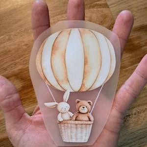 Könnte beinhalten: Eine skurrile Illustration eines Heißluftballons mit einem Hasen und einem Teddybären in einem Weidenkorb. Der Ballon ist cremefarben und hellbraun, mit dem Korb und den Tieren in weichen, gedämpften Tönen. Das Bild wird in einer Hand gehalten.
