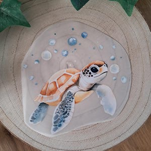 Könnte beinhalten: Ein Aquarellgemälde einer Meeresschildkröte mit orange-braunem Panzer, blauen Flossen und einem weißen Kopf. Die Schildkröte ist von blauen Blasen auf einem hellbeigen Hintergrund umgeben, der auf einer Holzscheibe platziert ist. Grüne Blätter sind im Hintergrund.