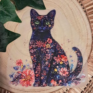 Könnte beinhalten: Ein Aufkleber mit einer schwarzen Katze mit grünen Augen, verziert mit bunten Blumenmustern. Die Katze sitzt, umgeben von Aquarellblumen in Rosa, Rot und Blau. Der Aufkleber ist auf einer Holzscheibe platziert.
