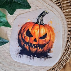 Könnte beinhalten: Aquarellillustration eines grinsenden Kürbisses für Halloween. Der Kürbis ist orange mit schwarzen Augen und einem Stiel. Spritzer verstärken das gruselige Thema. Das Bild befindet sich auf einem hellen, abgerundeten Quadrat.