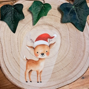 Könnte beinhalten: Aquarell-Illustration eines Rentiers mit Weihnachtsmütze, platziert auf einer Holzscheibe. Das Rentier hat braunes Fell, ein Geweih und rosige Wangen. Grüne Efeublätter sind um die Holzscheibe angeordnet und schaffen ein festliches Urlaubsthema.