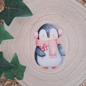 Könnte beinhalten: Eine Illustration eines Pinguins mit rosa Ohrenschützern und einem passenden Schal, der einen rot-weiß gewirbelten Lutscher hält. Der Pinguin ist vor einem hellen Hintergrund platziert und schafft eine charmante, winterliche Szene.