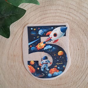 Könnte beinhalten: Eine flache, ausgestanzte Dekoration in Form der Zahl fünf mit einem dunkelblauen Weltraum-Motiv. Das Design zeigt eine weiße Rakete mit orangefarbenen Flammen, einen schwebenden Astronauten im weißen Anzug, bunte Planeten und kleine Sterne.