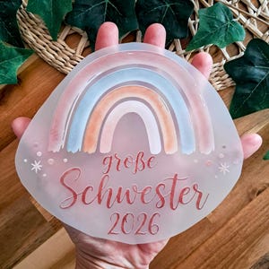 Könnte beinhalten: Ein klarer, ovaler Anhänger mit einem Aquarell-Regenbogen in Pastellfarben. Der Anhänger trägt den Schriftzug "große Schwester 2026" in roter Schrift. Kleine Sternformen sind um den Text herum verstreut.