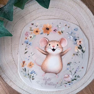 Könnte beinhalten: Eine Illustration einer lächelnden Maus mit ausgestreckten Armen, umgeben von bunten Blumen. Die Maus ist hellbraun mit rosa Ohren und einem langen Schwanz. Der Text "Hallo Frühling" steht unter der Maus.