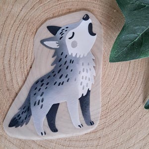 Könnte beinhalten: Ein skurriler Aufkleber mit einer Illustration eines heulenden Wolfs. Der Wolf ist in Grau- und Weißtönen gehalten, mit einem stilisierten Design. Der Aufkleber befindet sich auf einem hellen, unregelmäßig geformten Hintergrund, der an eine Holzscheibe erinnert.