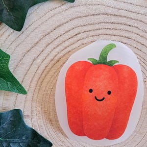 Könnte beinhalten: Ein roter Paprika-Aufkleber mit einem lächelnden Gesicht und einem grünen Stiel. Der Aufkleber befindet sich auf einer hellen Holzoberfläche mit grünen Blättern im Hintergrund.