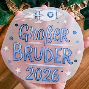 Könnte beinhalten: Ein transluzentes, rundes Schild mit dem Text "Großer Bruder 2026" in Blau, zusammen mit einer Rakete und Sternen. Das Schild wird vor einem Hintergrund aus Grün und Holz gehalten.