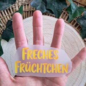 Könnte beinhalten: Ein durchscheinender Aufkleber mit den Worten "FRECHES FRÜCHTCHEN" in gelben und orangenen Buchstaben. Der Aufkleber wird in einer Hand gehalten, mit einem Holz-Hintergrund und grünen Blättern.