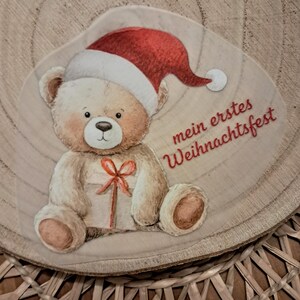 Könnte beinhalten: Rundes Holzornament mit einem Teddybär, der einen roten Weihnachtsmannhut trägt und ein Geschenk hält. Der Text "mein erstes Weihnachtsfest" ist in Rot geschrieben. Das Ornament hat eine natürliche Holzmaserung.