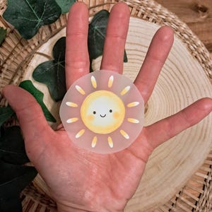 Könnte beinhalten: Ein runder, transparenter Aufkleber mit einer lächelnden Sonne mit gelben und orangefarbenen Strahlen. Die Sonne hat eine gelbe Mitte und ein fröhliches Gesicht. Der Aufkleber wird in einer Hand gehalten, mit einem Holz- und grünen Blätterhintergrund.