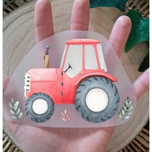 Könnte beinhalten: Illustration eines roten Traktors mit schwarzen Reifen und weißen Felgen. Der Traktor hat eine weiße Kabine und einen kleinen Schornstein. Die Illustration befindet sich auf einer klaren, abgerundeten Oberfläche mit Aquarellblättern.