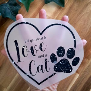 Könnte beinhalten: Ein herzförmiges Design mit den Worten "All you need is Love and a Cat" in schwarzer Schrift. Auf der rechten Seite befindet sich ein schwarzer Katzenpfotenabdruck. Das Design wird von einer Hand gehalten, mit einem Holz Hintergrund.
