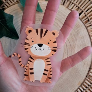 Könnte beinhalten: Ein transparenter Aufkleber mit einem Zeichentricktiger mit orangefarbenen und schwarzen Streifen. Der Tiger hat ein lächelndes Gesicht mit Schnurrhaaren und sitzt aufrecht. Der Aufkleber wird in einer Hand gehalten.