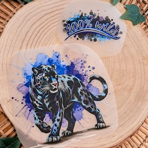Könnte beinhalten: Zwei runde, durchscheinende Aufkleber. Einer zeigt einen schwarzen Panther mit blauen und lila Aquarellakzenten. Der andere hat den Text "100% wild" in blauer Schrift. Beide sind vor einem Holzscheiben-Hintergrund platziert.