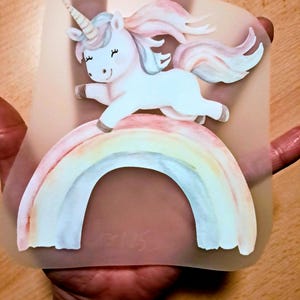Könnte beinhalten: Eine skurrile Illustration eines weißen Einhorns mit pastellfarbener Regenbogenmähne und -schwanz, das über einen Regenbogen springt. Das Einhorn hat ein spiralförmiges Horn und geschlossene Augen. Das Bild befindet sich auf einem durchscheinenden Hintergrund.