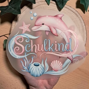 Könnte beinhalten: Ein Design mit einem rosa Delfin, Seesternen und Muscheln, mit dem Wort "Schulkind" in Schreibschrift. Das Design befindet sich auf einer transparenten, abgerundeten Oberfläche. Das Design hat blaue und rosa Akzente.