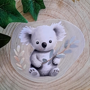 Könnte beinhalten: Niedliche Illustration eines Koala-Bären in Grau- und Weißtönen, der einen Zweig mit Blättern hält. Das Bild befindet sich auf einem hellen Holzmaserungshintergrund mit grünen Blättern in den Ecken.