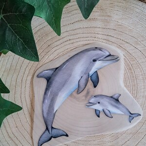 Könnte beinhalten: Eine Holzplakette mit einem Aquarellgemälde von zwei Delfinen. Der größere Delfin befindet sich über einem kleineren, beide in Grau- und Blautönen. Die Plakette befindet sich auf einer hellen Holzoberfläche.